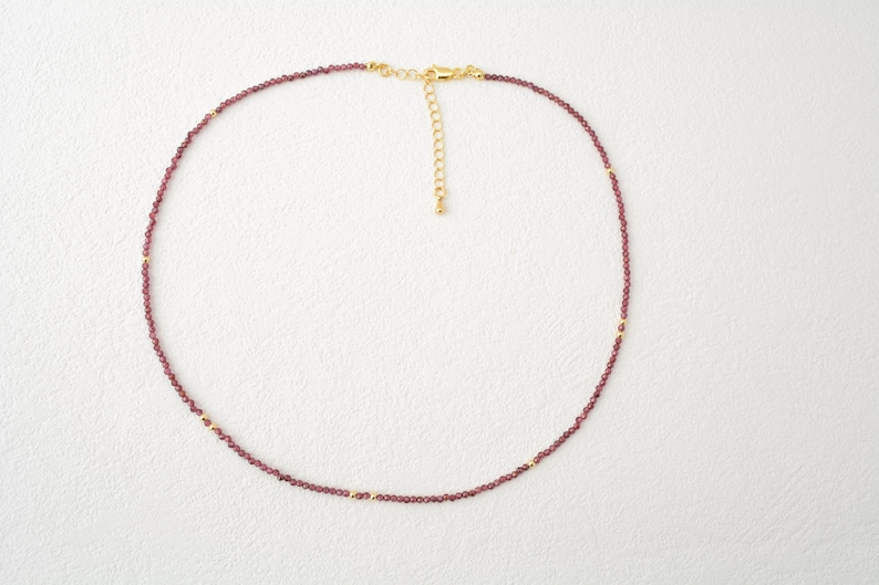 May include: A delicate necklace featuring small, deep red garnet beads strung together, accented with tiny gold-toned metal spacers. The piece includes a gold-toned lobster claw clasp and an adjustable extender chain for a customizable fit.