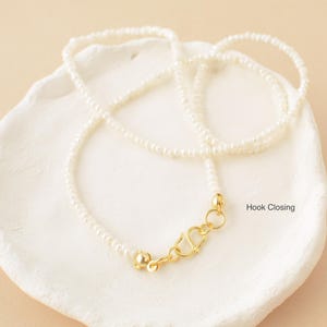 May include: A delicate white pearl necklace with a gold hook closure. The necklace is displayed on a white ceramic dish.
