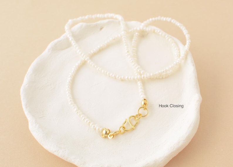 May include: A delicate white pearl necklace with a gold hook closure. The necklace is displayed on a white ceramic dish.