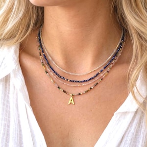 May include: A layered necklace set consisting of four strands of small, faceted gemstone beads in shades of clear, deep blue, light pink, and multicolored earth tones, featuring a single gold-toned letter A pendant on the lowest strand.