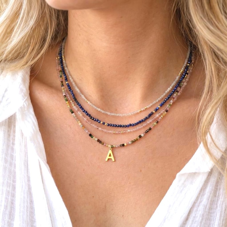 May include: A layered necklace set consisting of four strands of small, faceted gemstone beads in shades of clear, deep blue, light pink, and multicolored earth tones, featuring a single gold-toned letter A pendant on the lowest strand.