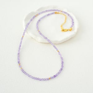 May include: A delicate necklace made with small, light purple beads and gold accents. The necklace is displayed on a white surface.