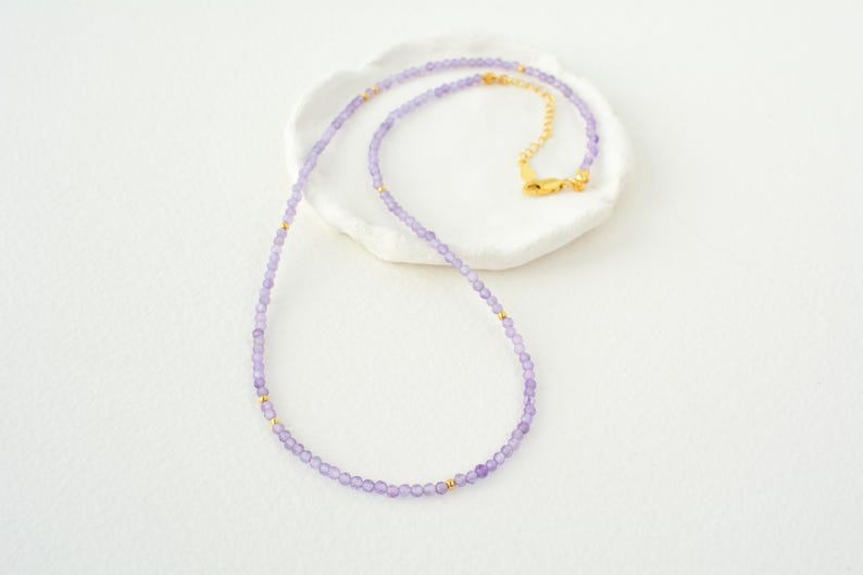 May include: A delicate necklace made with small, light purple beads and gold accents. The necklace is displayed on a white surface.