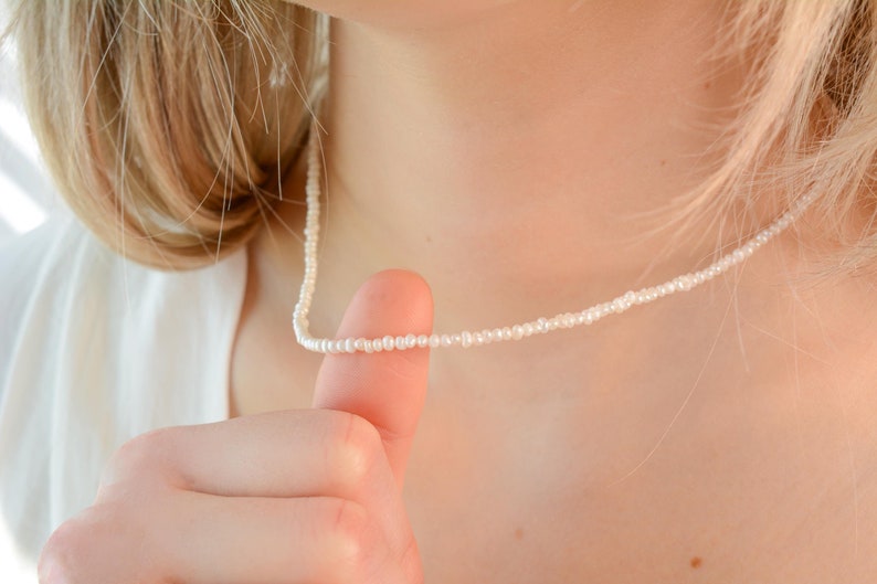 May include: A close-up of a woman wearing a simple white pearl necklace. The necklace is made of small, white pearls and is worn around the neck.