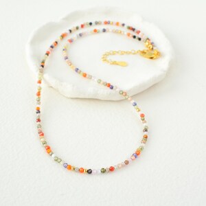 Puede incluir: Un delicado collar hecho de peque&ntilde;as cuentas de colores. Las cuentas son una mezcla de naranja, rojo, verde, azul y morado. El collar est&aacute; sobre una superficie blanca.