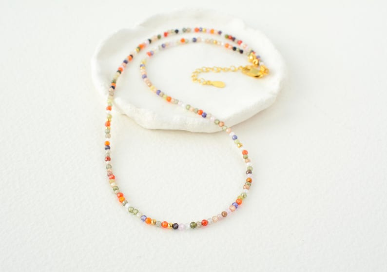 Puede incluir: Un delicado collar hecho de peque&ntilde;as cuentas de colores. Las cuentas son una mezcla de naranja, rojo, verde, azul y morado. El collar est&aacute; sobre una superficie blanca.