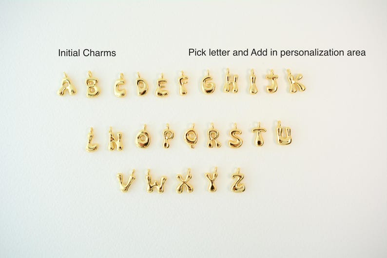 May include: Gold-colored initial charms arranged on a white surface. The image includes the alphabet from A to Z, with the text "Initial Charms" and "Pick letter and Add in personalization area".