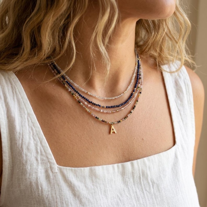 May include: A layered beaded necklace set featuring four strands of small, multi-colored faceted beads in shades of blue, pink, and clear. The lowest strand includes a small, gold-toned metal pendant in the shape of the capital letter A.