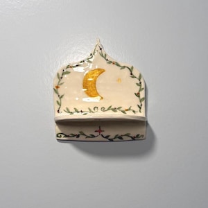 May include: A ceramic wall shelf with a cream-colored base, featuring a gold crescent moon and star design. The shelf is adorned with green and red painted vines. The shelf has a small ledge for displaying small items.