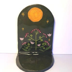 May include: A dark green, arched ceramic shelf with a painted design. The design includes a yellow sun, pink flowers, and stars. The shelf has a small ledge at the bottom.