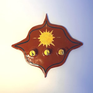 May include: A decorative wall-mounted key holder. The holder is a reddish-brown color with a gold sun design and three gold hooks. The edges are outlined in dark blue. The key holder is approximately 10 inches wide and 8 inches tall.