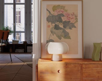Può includere: Una lampada da tavolo bianca a forma di fungo è appoggiata su un mobile in legno, accanto a una stampa botanica incorniciata con fiori rosa e foglie verdi. A destra c'è un vaso decorativo verde ondulato.