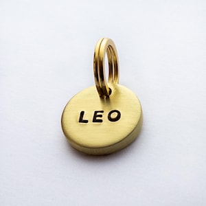 May include: Gold-coloured metal dog tag with a round shape and a split ring. The tag has the name "LEO" engraved in black lettering.