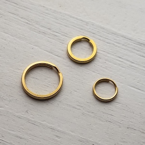 May include: Three gold-colored metal key rings of different sizes. The key rings are on a white wooden surface.