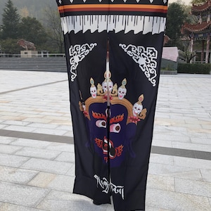 May include: Black fabric door curtain with a striking illustration of a deity, adorned with a skull crown and a stern expression. The top features a decorative border with white, orange, and brown accents. White script is at the bottom.