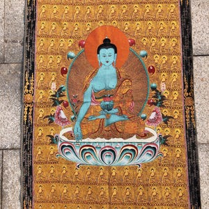 Op de afbeelding: Een gedetailleerde tapisserie met een zittende Boeddha met blauwe huid, omringd door een oranje halo. De Boeddha zit op een lotusbloem, met een achtergrond van herhaalde Boeddha-figuren in goud. De rand is zwart met gouden accenten.