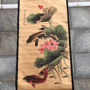 May include: A traditional Asian scroll painting featuring koi fish swimming among lotus flowers and leaves. The artwork uses a muted color palette with shades of brown, green, pink, and red. The scroll is framed with a black border.