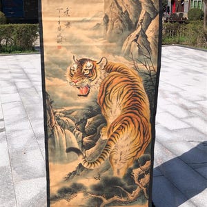 May include: A traditional Asian scroll painting featuring a detailed tiger with orange and black stripes, set against a mountainous landscape with a waterfall. The artwork is rendered in warm, earthy tones, with Chinese characters in the upper left corner.