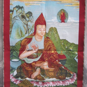 Puede incluir: Un tapiz que representa una figura sentada con túnicas rojas y doradas, con un gorro puntiagudo. La figura está rodeada por un paisaje de montañas y nubes, con un borde rojo y escritura tibetana en la parte inferior. La combinación de colores es rica y vibrante.