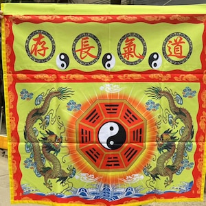 May include: A rectangular yellow banner with red and yellow trim. The banner features Chinese characters, yin-yang symbols, and two golden dragons flanking a red octagon with a yin-yang symbol in the center. The banner is held up by two hands.