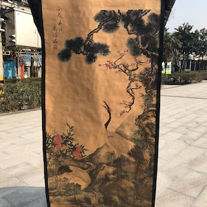 Puede incluir: Pintura tradicional en rollo asiático que representa dos grullas en un paisaje natural. La obra de arte muestra una grulla con el pico abierto, sobre un fondo de árboles, flores y montañas. El rollo está enmarcado con un borde negro.
