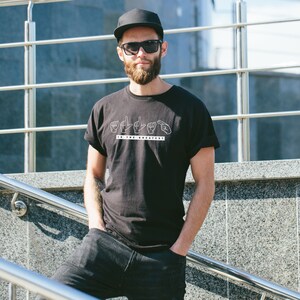 May include: A man wearing a black t-shirt with a white graphic of hands signing "I the greatest". He is also wearing a black baseball cap and sunglasses.