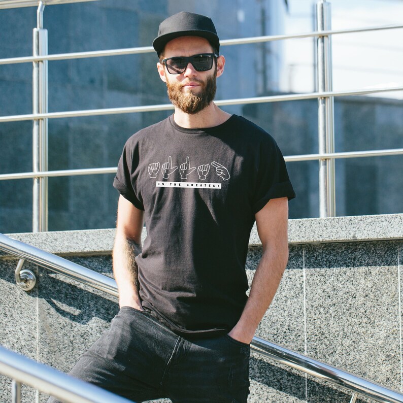 May include: A man wearing a black t-shirt with a white graphic of hands signing "I the greatest". He is also wearing a black baseball cap and sunglasses.
