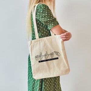 May include: A beige tote bag with black handles and the text "LOVE IS THE GREATEST" in black lettering. The text is in British Sign Language.