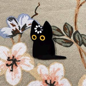 May include: A black cat enamel pin featuring large yellow circular eyes and a white loading icon symbol centered on the forehead. The cat is depicted in a sitting position with its tail extended to the right, rendered in a smooth, glossy finish.