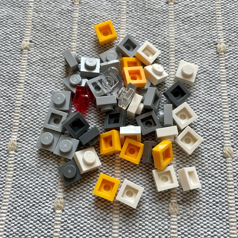 K&ouml;nnte beinhalten: Eine Sammlung kleiner Plastikbausteine in verschiedenen Farben, darunter Grau, Wei&szlig; und Gelb. Die Steine sind auf einer grau-wei&szlig; gestreiften Oberfl&auml;che zu einem Haufen gestapelt.