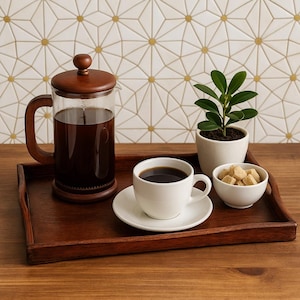 May include: A wooden serving tray holds a French press coffee maker, a white coffee cup and saucer, a small bowl of sugar cubes, and a potted plant. The coffee is dark, and the tray is set against a geometric tile background.