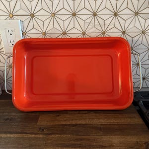 May include: A vibrant red rectangular baking tray with silver handles. The tray is empty and rests on a wooden surface. The background features a white and gold geometric tile pattern.