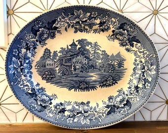 Op de afbeelding: Ovale blauwe en witte keramische schaal met een gedetailleerde landschapsscène in het midden. De rand heeft bloemen- en bladerenontwerpen. De schaal staat op een houten oppervlak tegen een achtergrond van tegels met patronen.