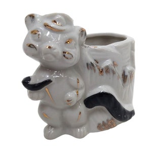 May include: White ceramic planter shaped like a squirrel holding a pipe. The planter has gold accents and a black tail. The planter is sitting on a white background.