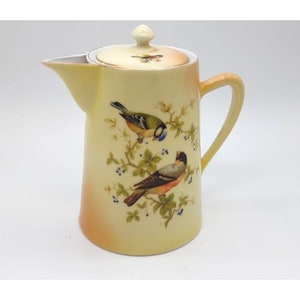 May include: A yellow ceramic creamer with a lid, featuring a painted design of two birds perched on branches with green leaves and blue berries.