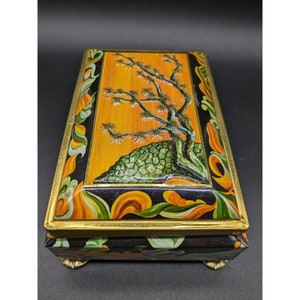May include: A vintage tin box with a black background and gold trim. The lid features a painted scene of a stylized tree with green foliage on a yellow background. The box has four gold feet.