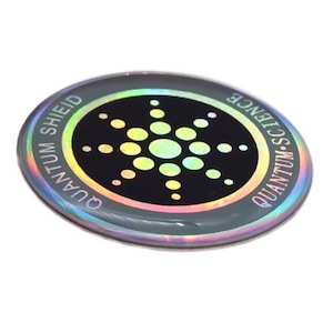 Pode incluir: Um autocolante oval holográfico com as palavras "QUANTUM SHIELD" e "QUANTUM SCIENCE" na borda. O centro apresenta um círculo preto com um design abstrato colorido. O autocolante tem um acabamento refletivo e iridescente.