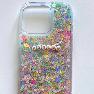 May include: Clear phone case with a glittery design featuring a variety of colorful flowers, stars, and other shapes. The name "ASHLEE" is spelled out in white letters with gold outlines.