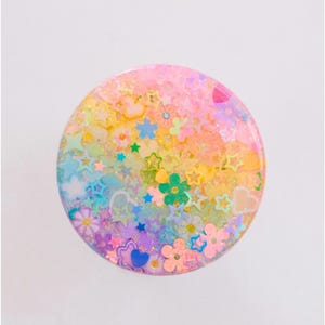 May include: A round, colorful phone grip with a glittery, multi-colored design. The grip features various shapes, including stars, flowers, and hearts, in shades of pink, yellow, blue, and green. The overall aesthetic is bright and playful.