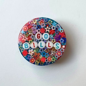 May include: A round, colorful phone grip with a glittery background and the words "GO BILLS" spelled out in white letters with red and blue accents.