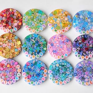 May include: Twelve circular resin coasters featuring embedded colorful glitter, small flower and star shapes, and white alphabet beads spelling out the twelve zodiac signs: Aries, Taurus, Gemini, Cancer, Leo, Virgo, Libra, Scorpio, Sagittarius, Capricorn, Aquarius, and Pisces.