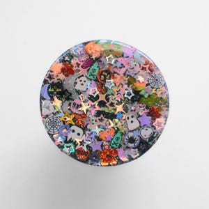 May include: A round, glittery Halloween-themed object with a variety of small, colorful shapes embedded within. The shapes include stars, ghosts, spiders, pumpkins, and spiderwebs in orange, purple, green, and black.