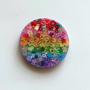 May include: A round, colorful resin piece with a rainbow of small, decorative elements. The design includes flowers, hearts, and fruit shapes, with glitter accents. The piece is set against a white background.