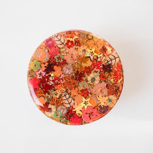 May include: A round, clear resin object filled with autumn-themed embellishments. The design includes miniature pumpkins, leaves, flowers, and stars in shades of orange, red, and gold, with glitter accents. The overall effect is festive and decorative.