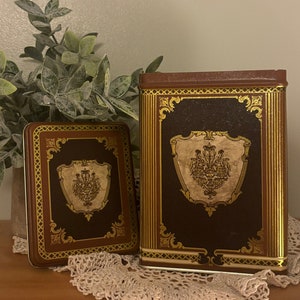 May include: Two decorative boxes with a brown background and gold trim. The boxes have a crest design with gold accents. The larger box has a crest with a crown and the smaller box has a crest with a crown and a heart.