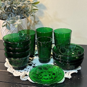May include: A collection of green glass tableware, including bowls, glasses, and a saucer, arranged on a white lace doily. The glassware features a detailed embossed pattern. A potted plant is visible in the background, adding a touch of natural decor.
