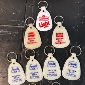 May include: A collection of vintage Schmidt beer keychains. The keychains are white with a rounded bottom and a metal ring. Some keychains have the text "Light Beer" in red. Other keychains have the text "Nelson Beer Distributor" in red or blue.