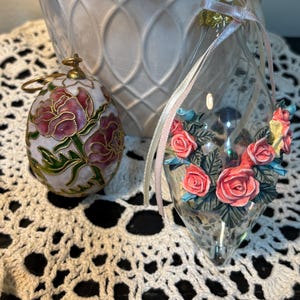 May include: Two decorative ornaments. One is egg-shaped with pink roses and gold accents. The other is a clear, teardrop-shaped glass ornament adorned with pink roses and blue accents. Both have ribbons for hanging.