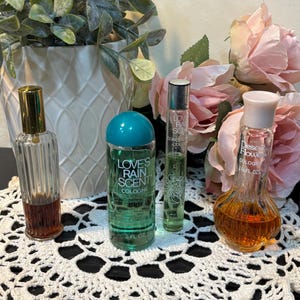 May include: An assortment of vintage perfume bottles, including a bottle with a gold cap and a bottle labeled "Love's Rain Scent Cologne". The bottles are arranged on a white lace doily, with pink roses and greenery in the background.