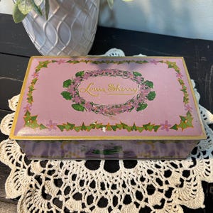 May include: A rectangular, vintage Louis Sherry tin box. The box is a soft lavender color with a gold trim. The lid features a floral wreath design with the brand name in gold script. The box is resting on a white lace doily.